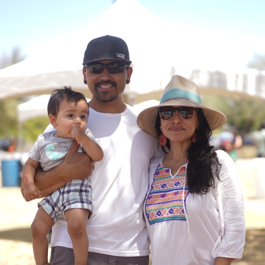 Young child in plaid shorts held by both parents. The father is wearing a baseball cap. the mother is wearing a straw hat.