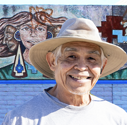 Smiling elderly gentleman with a sun hat in front of El Pueblo mural